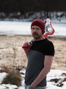 Thomas Franz steht mit einer Spielzeuggitarre an einem verschneiten Fluss